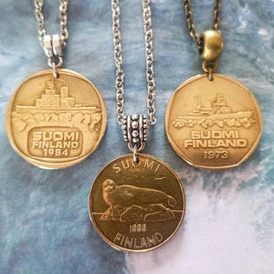 May include: Three gold-toned coin pendants on silver chains. The coins feature images of a ship and a seal, with the words "SUOMI FINLAND" and the years 1973, 1984, and 1993. The pendants are displayed against a blue background.