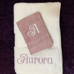May include: A white bath towel with the embroidered name "Aurora" in a script font, and a smaller mauve hand towel with a white embroidered "A". The towels are on a dark purple surface.