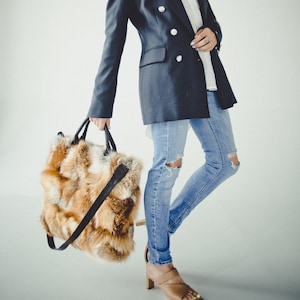May include: A woman wearing a dark blue blazer, white shirt, and blue jeans with distressed details. She is holding a brown and orange fur tote bag with a black leather strap.