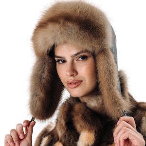 Puede incluir: Una mujer lleva un abrigo de piel marrón y blanco con un gorro de piel a juego. El gorro tiene orejeras y un ribete de cuero.