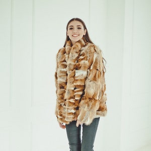 May include: A woman wearing a brown and white fur coat with a high collar, dark blue jeans, and white sneakers. She is standing in a white room with a white wall behind her.