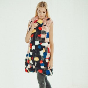 May include: A sleeveless faux fur vest in a patchwork design of black, pink, red, blue, yellow, and white. The vest is worn over grey jeans and white trainers. The model has blonde hair.