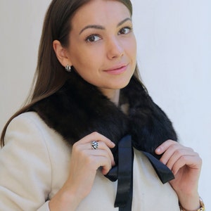 Puede incluir: Una mujer con un abrigo blanco con un cuello de piel negro y un lazo de sat&eacute;n negro. Lleva un anillo de plata en el dedo.
