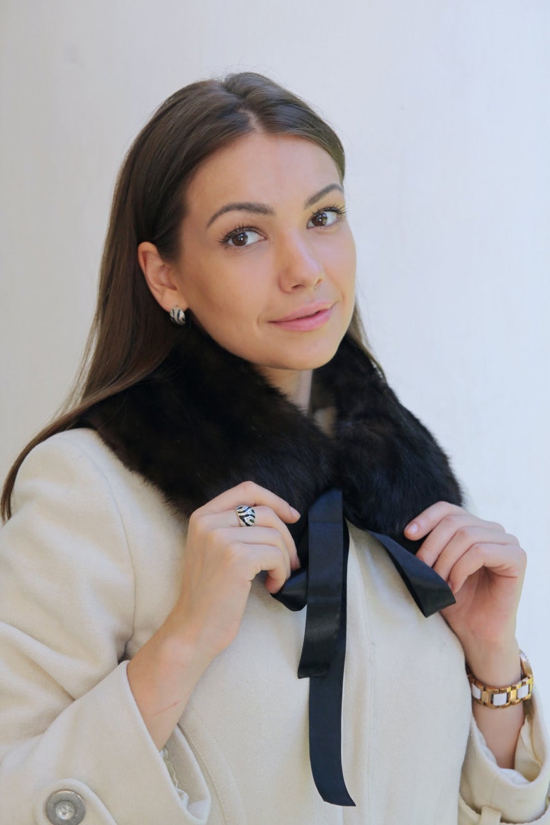 Puede incluir: Una mujer con un abrigo blanco con un cuello de piel negro y un lazo de sat&eacute;n negro. Lleva un anillo de plata en el dedo.