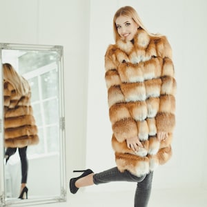May include: A woman wearing a long, orange and white fur coat. She is standing in front of a mirror and looking at herself.