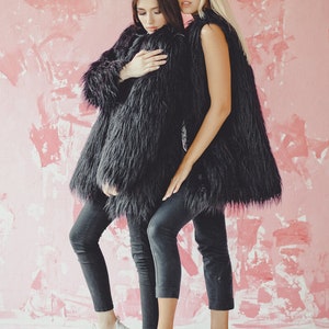 May include: Two people wearing black faux fur coats and dark gray jeans stand in front of a pink wall. One person wears a long coat, the other a vest. Both wear white sneakers. The image has the text "Florence photo" at the bottom.