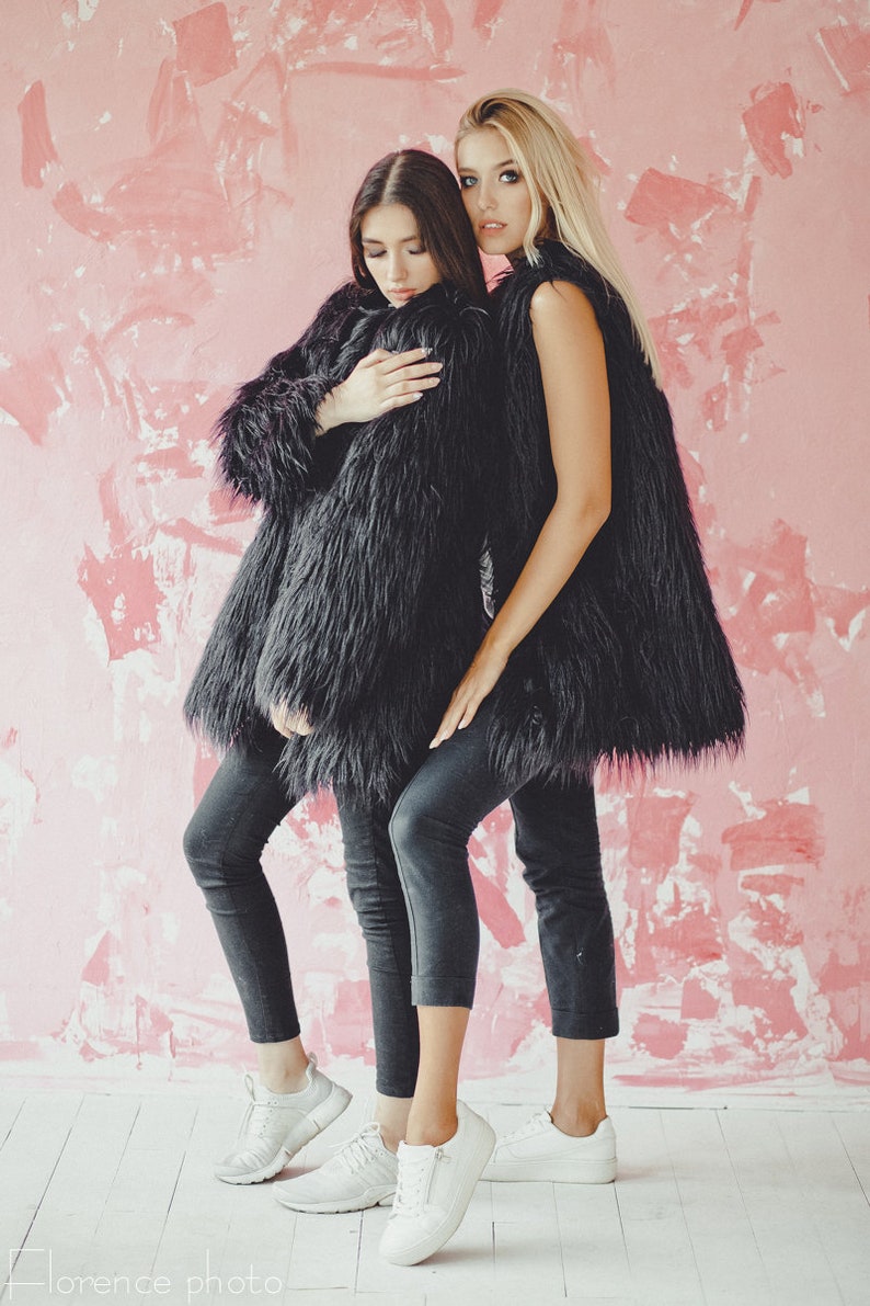 May include: Two people wearing black faux fur coats and dark gray jeans stand in front of a pink wall. One person wears a long coat, the other a vest. Both wear white sneakers. The image has the text "Florence photo" at the bottom.