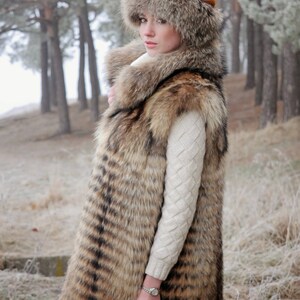 May include: A long, brown and black fur vest with a large fur collar. The vest is worn over a white sweater. The person is wearing a brown fur hat.