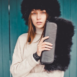 May include: A woman wearing a black fur hat and a cream sweater holds a black clutch purse. The background is a teal blue wall.