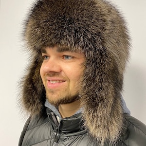 Pode incluir: Um chapéu de pele de trapper marrom com abas para os ouvidos. O chapéu é feito de pele verdadeira e tem um forro grosso e quente. O chapéu é usado por uma pessoa com barba.