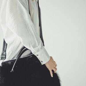 May include: A woman wearing a white shirt carries a black furry handbag with a long strap.