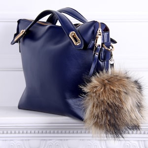 May include: A dark blue leather handbag with a gold zipper and hardware. The bag has a fluffy brown and black fur pom-pom attached to the zipper pull.