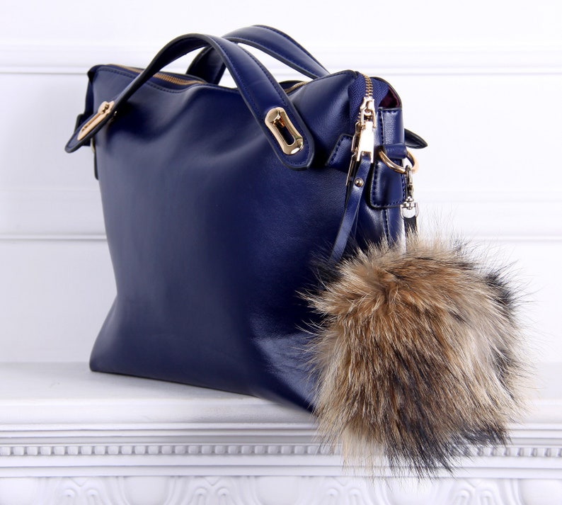 May include: A dark blue leather handbag with a gold zipper and hardware. The bag has a fluffy brown and black fur pom-pom attached to the zipper pull.