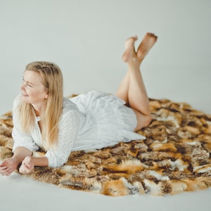 May include: A woman in a white dress is lying on a round, brown and white faux fur rug. She is smiling and looking to the side.
