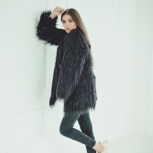 May include: A woman wearing a long, dark gray faux fur coat and dark gray leggings. She is also wearing white sneakers. The coat has long sleeves and a shaggy texture. The background is a plain white wall.