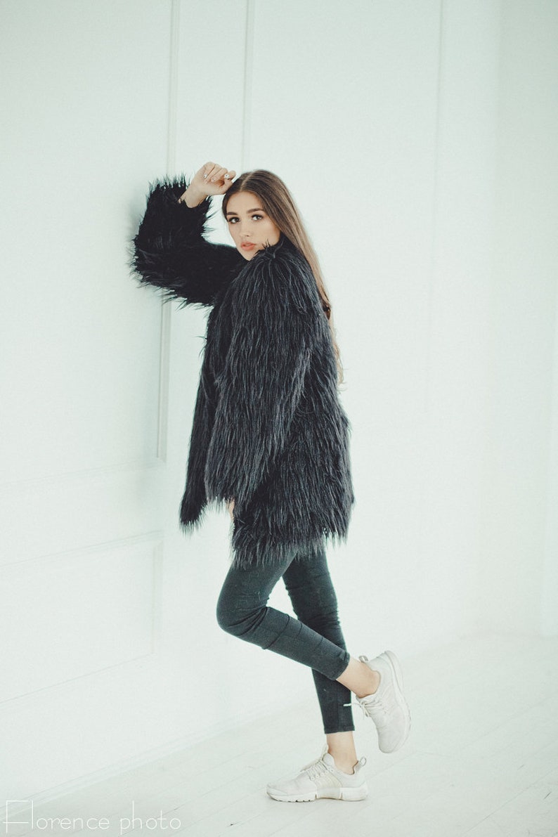 May include: A woman wearing a long, dark gray faux fur coat and dark gray leggings. She is also wearing white sneakers. The coat has long sleeves and a shaggy texture. The background is a plain white wall.