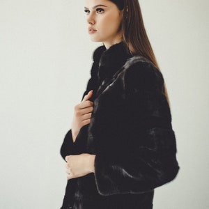 K&ouml;nnte beinhalten: Eine Frau tr&auml;gt eine schwarze Pelzjacke mit hohem Kragen und blauen Jeans. Die Jacke hat einen Knopfverschluss und ein elegantes, modernes Design.