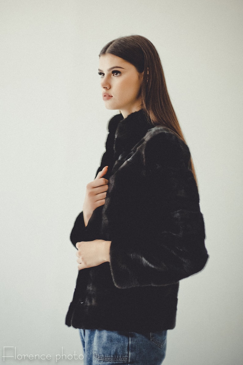K&ouml;nnte beinhalten: Eine Frau tr&auml;gt eine schwarze Pelzjacke mit hohem Kragen und blauen Jeans. Die Jacke hat einen Knopfverschluss und ein elegantes, modernes Design.