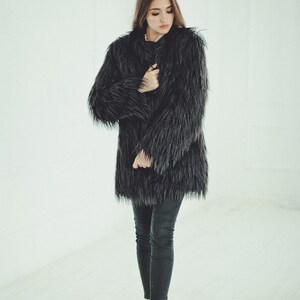May include: A long, black faux fur coat with a textured appearance. The coat is worn with black pants and white sneakers. The image is set against a bright, white background.