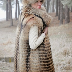 May include: A long, striped fur vest with a matching fur hat. The vest is brown and black with a white knit sweater underneath. The model is standing in a snowy forest.