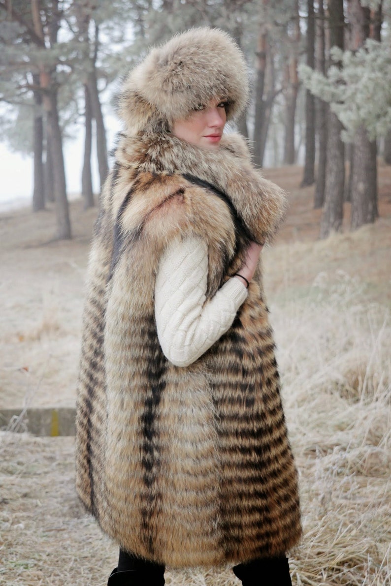 May include: A long, striped fur vest with a matching fur hat. The vest is brown and black with a white knit sweater underneath. The model is standing in a snowy forest.