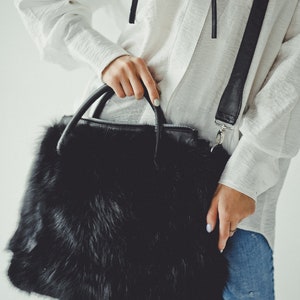 May include: A black faux fur handbag with a long black strap and silver hardware. The bag is being held by a person wearing a white shirt and blue jeans.