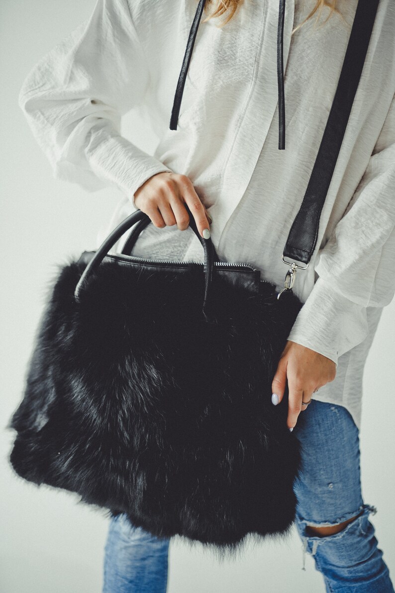 May include: A black faux fur handbag with a long black strap and silver hardware. The bag is being held by a person wearing a white shirt and blue jeans.
