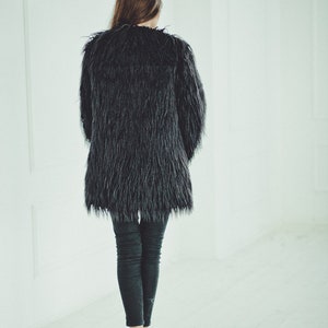May include: A black faux fur coat with a long, shaggy texture. The coat is worn over dark gray leggings and white sneakers. The person is walking away from the camera in a bright, white-walled space.