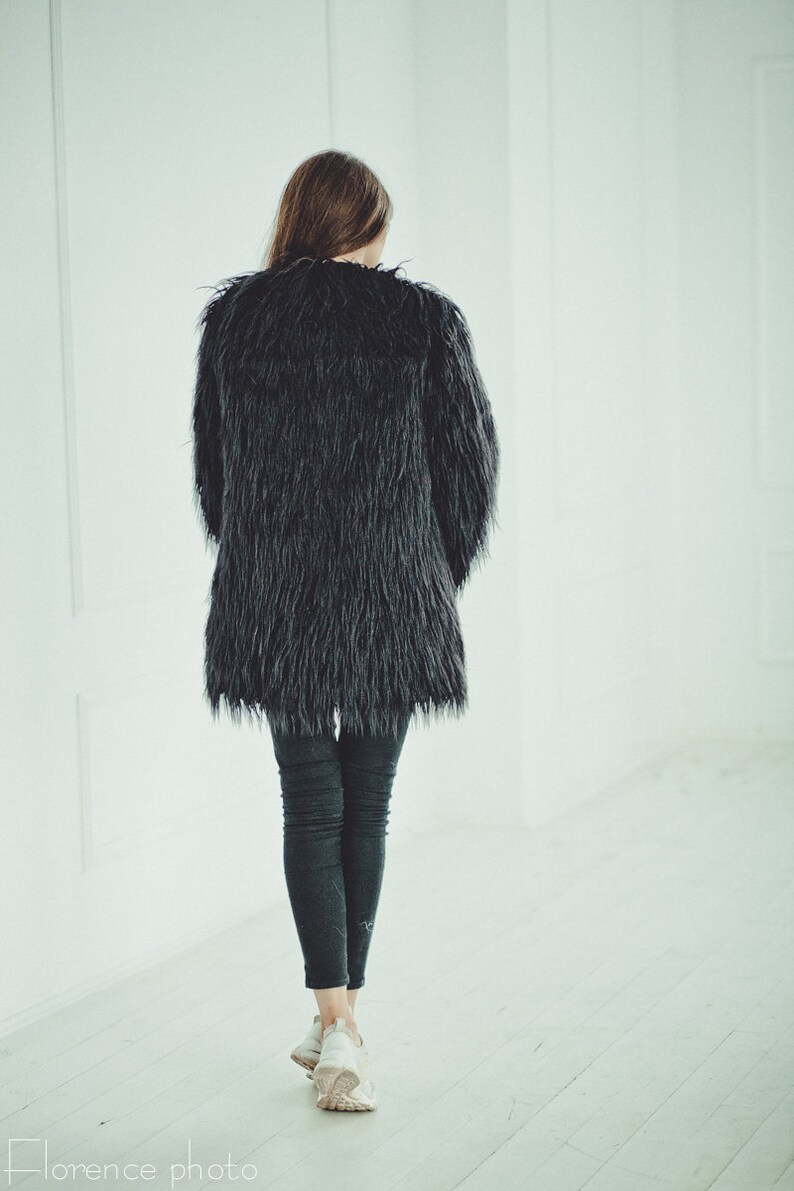 May include: A black faux fur coat with a long, shaggy texture. The coat is worn over dark gray leggings and white sneakers. The person is walking away from the camera in a bright, white-walled space.