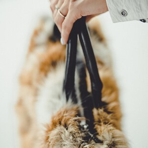 May include: A black leather handbag with a long strap and a furry brown and white lining. The bag is being held by a hand with a silver ring.