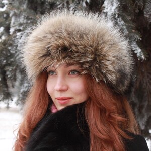 May include: A woman with long, red hair is wearing a brown fur hat with a wide brim. She is wearing a black coat and has a soft, natural look.