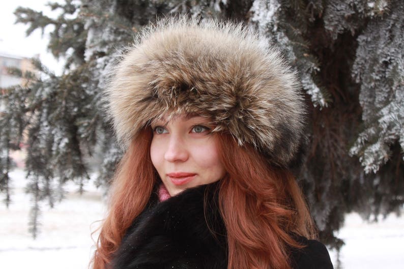 May include: A woman with long, red hair is wearing a brown fur hat with a wide brim. She is wearing a black coat and has a soft, natural look.