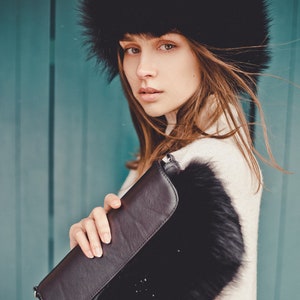 May include: A woman wearing a black fur hat and a white sweater holds a black clutch purse with a fur trim. The background is a teal blue wall.