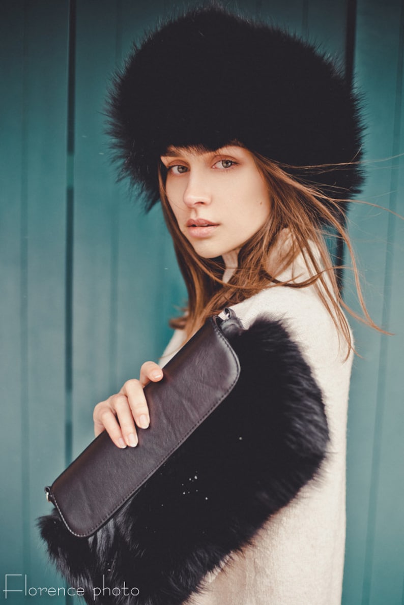 May include: A woman wearing a black fur hat and a white sweater holds a black clutch purse with a fur trim. The background is a teal blue wall.