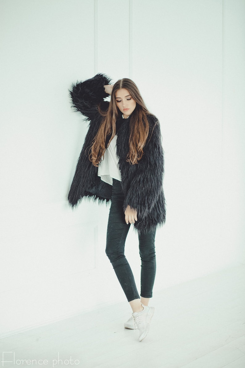 May include: A woman wearing a black faux fur coat, white top, and black pants. The coat has long, shaggy fur. She is wearing white sneakers. The background is a plain white wall.
