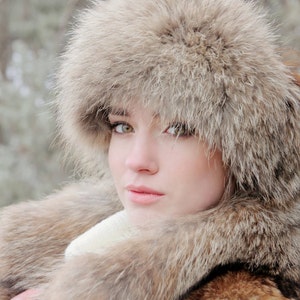 May include: A woman wearing a large, fluffy fur hat and matching fur coat. The hat covers most of her face, but her eyes are visible. The fur is a light brown color.