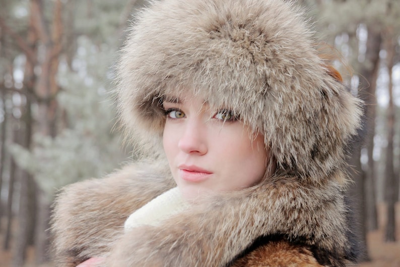 May include: A woman wearing a large, fluffy fur hat and matching fur coat. The hat covers most of her face, but her eyes are visible. The fur is a light brown color.