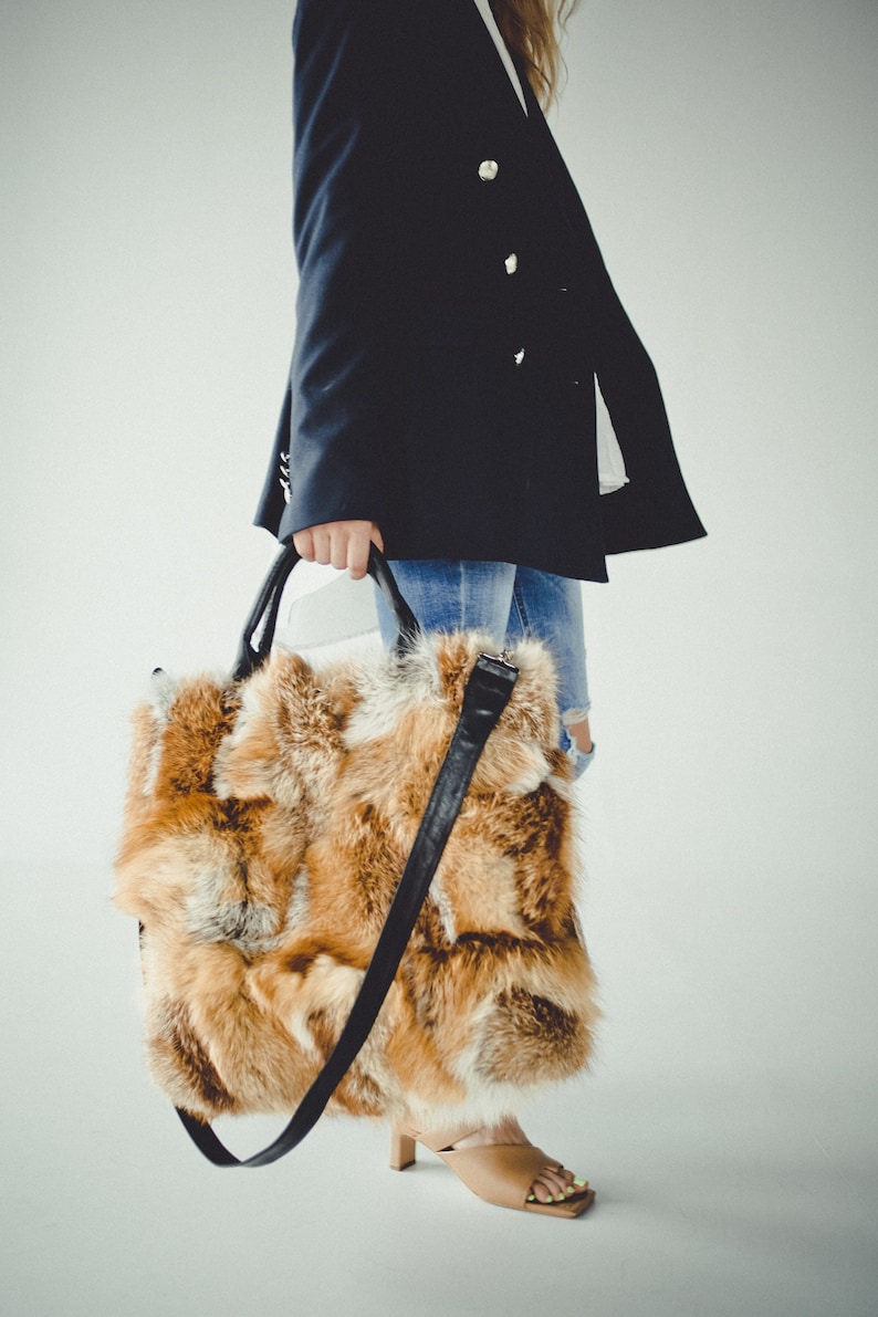 May include: A large, brown and white fur tote bag with a black leather strap. The bag is being held by a person wearing a dark blue blazer and jeans.