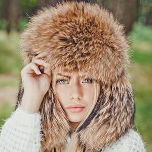 May include: A woman wearing a brown fur hat with a long, fluffy brim. The hat covers most of her face, but her eyes, nose, and mouth are visible. She is wearing a white sweater and has long, blonde hair.