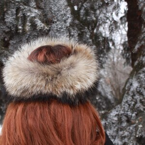 May include: A brown and white fur hat with a black band. The hat is worn by a person with long, red hair. The background is a snowy forest.