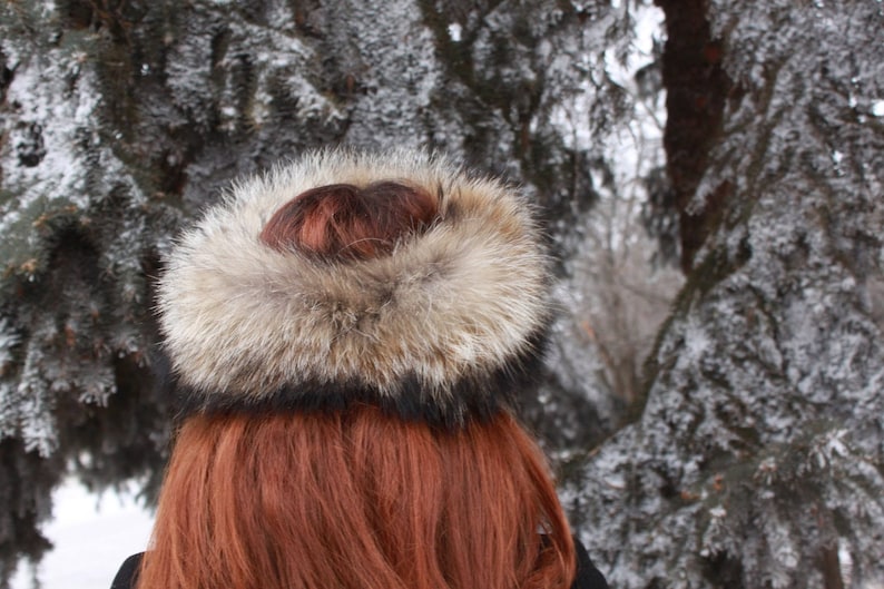 May include: A brown and white fur hat with a black band. The hat is worn by a person with long, red hair. The background is a snowy forest.