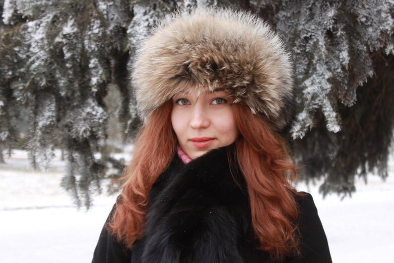May include: A woman wearing a brown fur hat with a black fur collar and a black coat. She is standing in front of a snowy background.