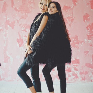 May include: Two people wearing black faux fur vests, black leggings, and white sneakers pose in front of a pink textured wall. One person has blonde hair, and the other has dark hair. The floor is white.