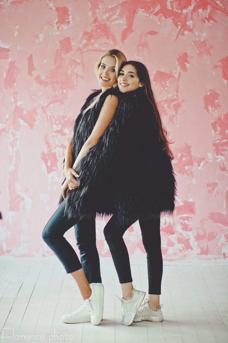May include: Two people wearing black faux fur vests, black leggings, and white sneakers pose in front of a pink textured wall. One person has blonde hair, and the other has dark hair. The floor is white.