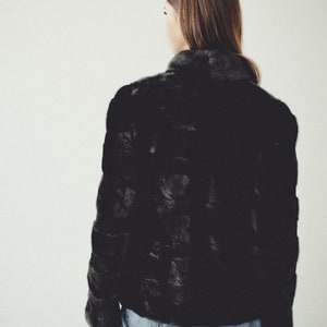 K&ouml;nnte beinhalten: Eine schwarze Pelzjacke mit hohem Kragen und langen &Auml;rmeln. Die Jacke wird &uuml;ber einer blauen Jeans getragen.