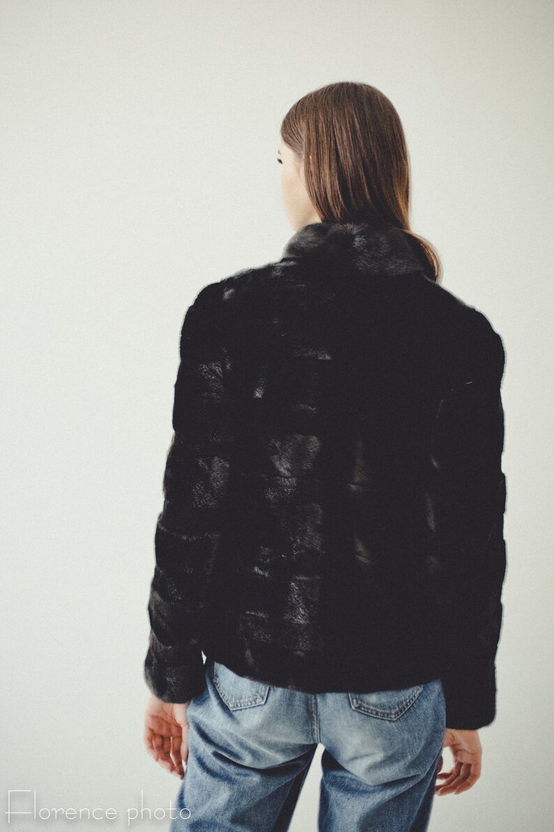 K&ouml;nnte beinhalten: Eine schwarze Pelzjacke mit hohem Kragen und langen &Auml;rmeln. Die Jacke wird &uuml;ber einer blauen Jeans getragen.