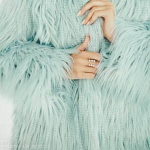 Puede incluir: Una mano de mujer con un anillo con varias piedras peque&ntilde;as, la mano es parcialmente visible a trav&eacute;s de una chaqueta de pelo azul claro.