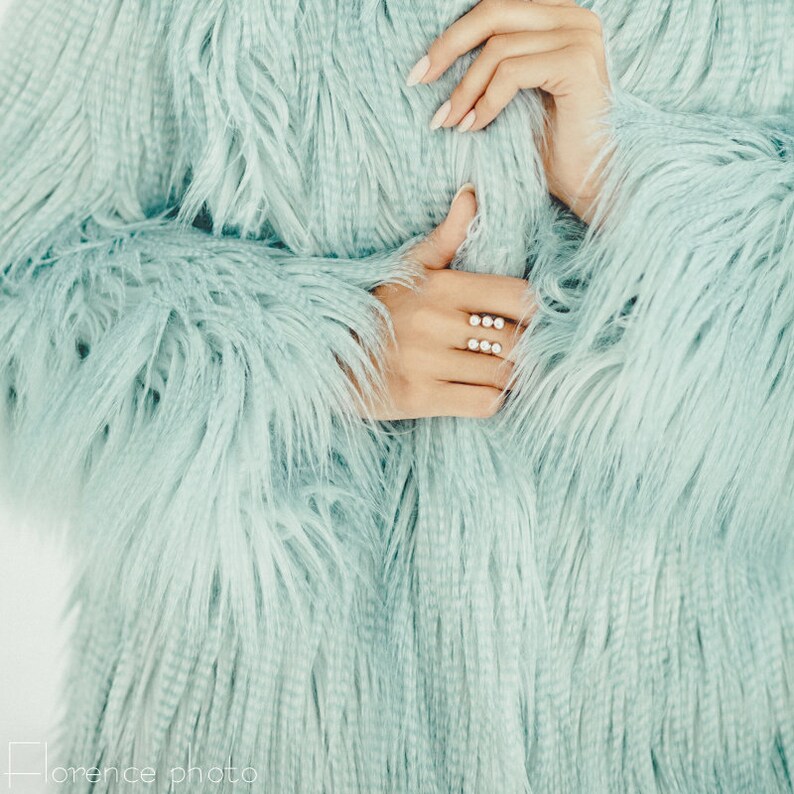 Puede incluir: Una mano de mujer con un anillo con varias piedras peque&ntilde;as, la mano es parcialmente visible a trav&eacute;s de una chaqueta de pelo azul claro.