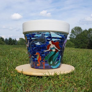 May include: A white ceramic planter decorated with a mosaic of colorful sea creatures, including a mermaid, seahorses, and a stingray. The mosaic is set against a blue background.