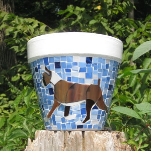 May include: A mosaic-tiled flower pot featuring a Boxer dog design. The pot has a white rim and is decorated with blue and white tiles. The Boxer is depicted in shades of brown, white, and black. The pot is displayed outdoors.